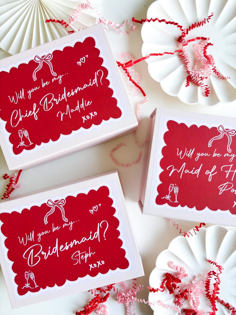 May include: Three pink gift boxes with red and white labels. The labels have a white scalloped border and say "Will you be my... Chief Bridesmaid? Maddie xoxo", "Will you be my... Bridesmaid? Steph xoxo", and "Will you be my Maid of Honor?"