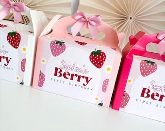 May include: Pink and white party favor boxes with strawberry designs and pink bows. The boxes feature the text "Sophia's Berry First Birthday" and are decorated with strawberries and daisies. The boxes have handles.