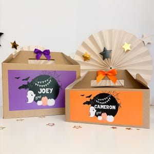 May include: Two brown cardboard gable boxes with handles. The larger box has a purple label with a black circle, a ghost, pumpkins, bats, and the name JOEY. The smaller box has an orange label with a black circle, a ghost, pumpkins, and CAMERON.