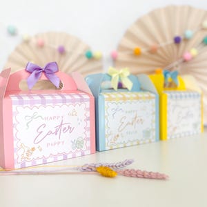 Peut inclure: Trois boîtes cadeaux de couleur pastel avec un motif à carreaux et un nœud sur le dessus. Les boîtes sont décorées d'un motif de Pâques et du texte "Joyeuses Pâques" et "Poppy".