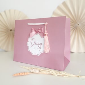 May include: A pink gift bag with a white handle and a pink bow. The bag has a white sticker with the text "Daisy Bride to Be" in black script.