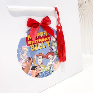 May include: White gift bag with a red bow and tassel. A circular graphic features characters from the Toy Story film and the text "HAPPY BIRTHDAY BILLY". The bag has a white handle.