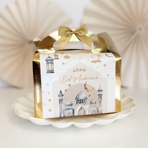 May include: A gold gift box with a white and blue design featuring a mosque, stars, and a crescent moon. The box has a gold bow and the text "Adam Eid Mubarak" on the front.