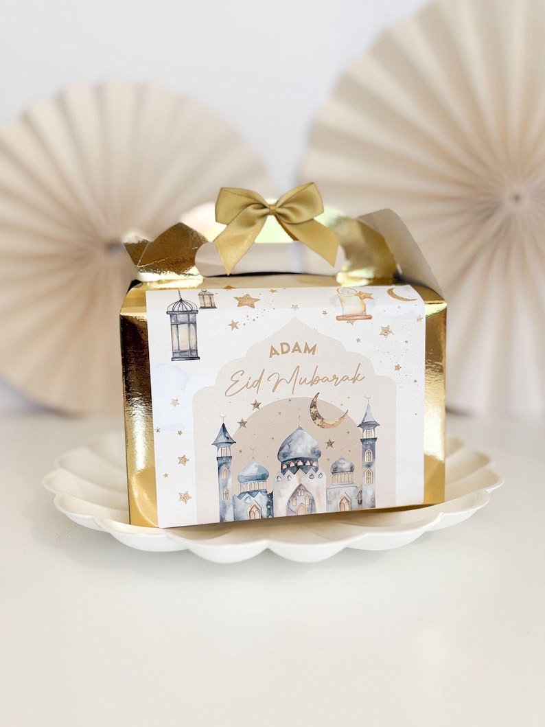 May include: A gold gift box with a white and blue design featuring a mosque, stars, and a crescent moon. The box has a gold bow and the text "Adam Eid Mubarak" on the front.