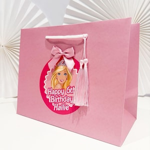 May include: A pink gift bag with a Barbie graphic and the text "Happy 6th Birthday Hallie." The bag is decorated with a pink bow and tassel. It has white cord handles. Ideal for a birthday party.