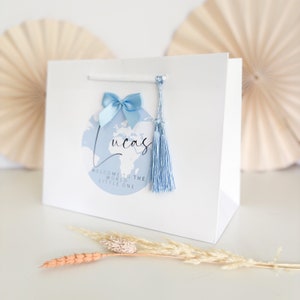 May include: White gift bag with a blue tassel and bow. The bag has a blue and white world map design with the text "Lucas" and "Welcome to the world little one".