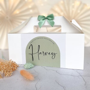 May include: White gift box with a handle and a sage green bow. The box has a label with the name "Harvey" in script font and the text "THANK YOU FOR BEING OUR FLOWER GIRL". Dried flowers are in the foreground.