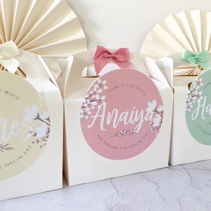 May include: Three white favour boxes with pink, green and yellow floral designs. The boxes have a handle and a circle sticker with the text "Thank you so much" and a name. The pink box says "Anaiya" and the green box says "Hailey".