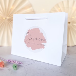 May include: White gift bag with a pink and white circle sticker that says "Jasmine Maid of Honour".