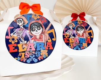 May include: White favor boxes with colorful cartoon characters and the text "Elara is 3" on a blue background. The boxes have red and white ribbon bows.