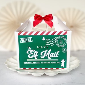 May include: A white gift box with a red bow. The box has a green panel with the text "LILY'S Elf Mail" and a red and white striped border. The box is on a white scalloped plate. The text includes the address: 25 Elf Lane, North Pole.