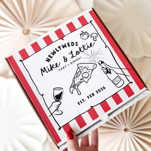 Può includere: Una scatola di pizza bianca con strisce rosse e bianche. La scatola ha un'illustrazione in bianco e nero di una fetta di pizza, un bicchiere di vino e una bottiglia di champagne. Il testo sulla scatola recita "Newlyweds Mike & Lottie That's amore! Est. Feb 2026".