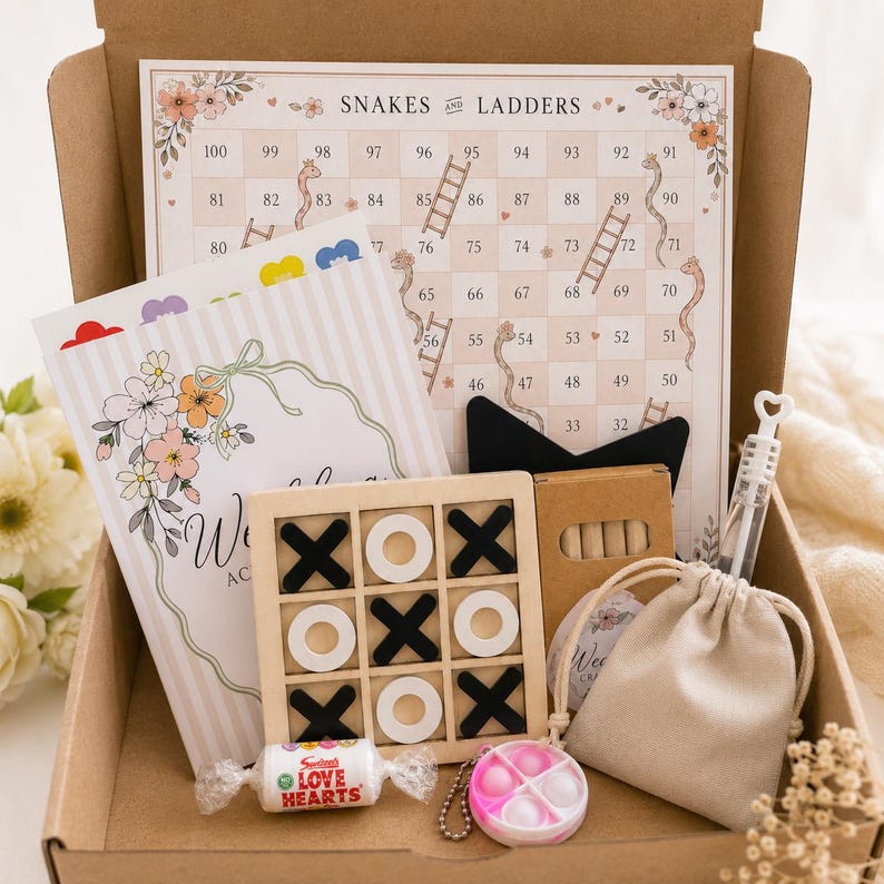 May include: A cardboard box containing a Snakes and Ladders game board, a floral activity booklet, a wooden tic-tac-toe set with black and white pieces, a small beige drawstring pouch, a bubble wand, a Love Hearts candy roll, and a small fidget toy.