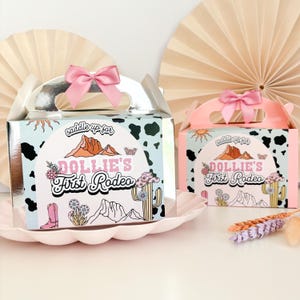 Peut inclure: Deux boîtes de faveur de fête argentées et roses avec un motif imprimé de vache. Les boîtes ont une poignée et un nœud rose. Les boîtes ont un autocollant avec le texte "saddle up for Dollie's First Rodeo".