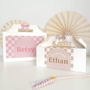May include: Two white gift boxes with pink and brown checkered patterns. The boxes have cut-out handles and are decorated with pink and brown ribbons. The boxes are labeled "Big Sister Betsy" and "Big Brother Ethan".