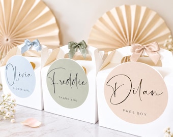 May include: Three white cardboard gable boxes with handles, each topped with a satin ribbon bow. The boxes feature circular labels in light blue, sage green, and beige, printed with the names Olivia, Freddie, and Dilan, respectively.