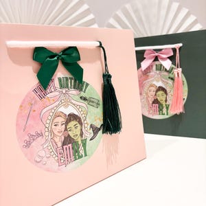 May include: Pink gift bag with a green bow and tassel. The bag features a circular graphic with two illustrated women, a crown, and the words "HAPPY BIRTHDAY EMILY". A second green and pink gift bag is visible in the background.