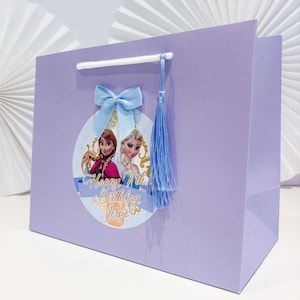 Puede incluir: Una bolsa de regalo lila con un gr&aacute;fico circular de Anna y Elsa, con el texto "Happy 7th Birthday Jersie". La bolsa tiene un lazo y una borla de color azul claro.