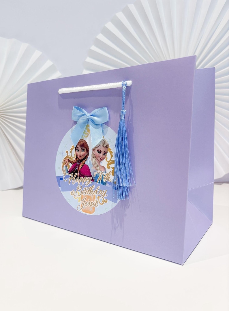 Puede incluir: Una bolsa de regalo lila con un gr&aacute;fico circular de Anna y Elsa, con el texto "Happy 7th Birthday Jersie". La bolsa tiene un lazo y una borla de color azul claro.