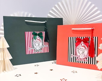 May include: Two Christmas gift bags, one dark green and one coral. Each bag has a rectangular label with a Santa Claus illustration, vertical stripes, and the text "Merry Christmas" and a name. The green bag has a green bow and tassel, the coral bag has a red bow and tassel.