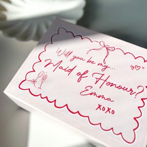 May include: A pink box with a red border and a white sticker that says "Will you be my Maid of Honour? Emma xoxo" with a drawing of two champagne glasses and a bow.