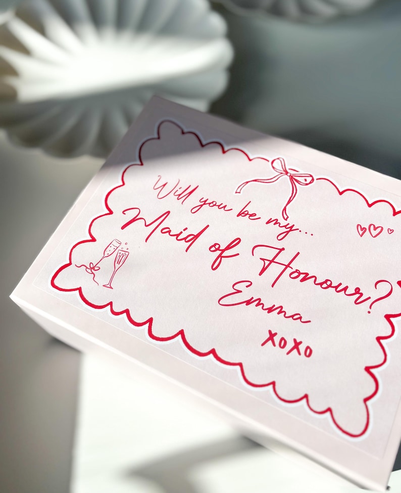 May include: A pink box with a red border and a white sticker that says "Will you be my Maid of Honour? Emma xoxo" with a drawing of two champagne glasses and a bow.
