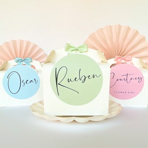 May include: Three white favour boxes with pastel coloured circles and names written in black script. The names are Oscar, Rueben, and Courtney. The box labelled Courtney also has the words "Flower Girl" written below the name.