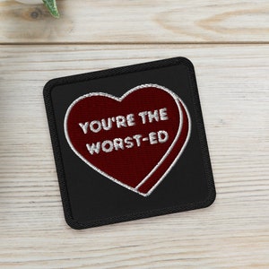 May include: A black square patch with a red heart design and the embroidered text "YOU'RE THE WORST-ED". Two small potted succulents are in the background on a light wood surface.