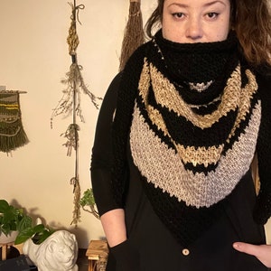 Brown haired woman in front of dried herbs with a sample of the Gabriel Tunisian crochet shawl pattern made using black, silver and gold single ply yarn, display the unique texture of the beginner friendly easy crochet stitch project.