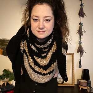 Brown haired woman in front of dried herbs with a sample of the Gabriel Tunisian crochet shawl pattern made using black, silver and gold single ply yarn, display the unique texture of the beginner friendly easy crochet stitch project.