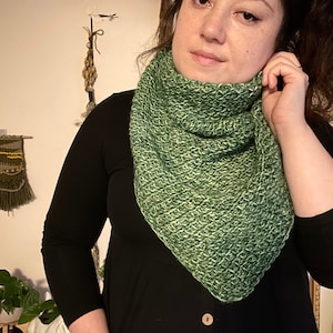 Brown haired woman in front of dried herbs with a sample of the Gabriel Tunisian crochet shawl pattern made using green ply yarn, display the unique texture of the beginner friendly easy crochet stitch project. Shown in kerchief size option reversed