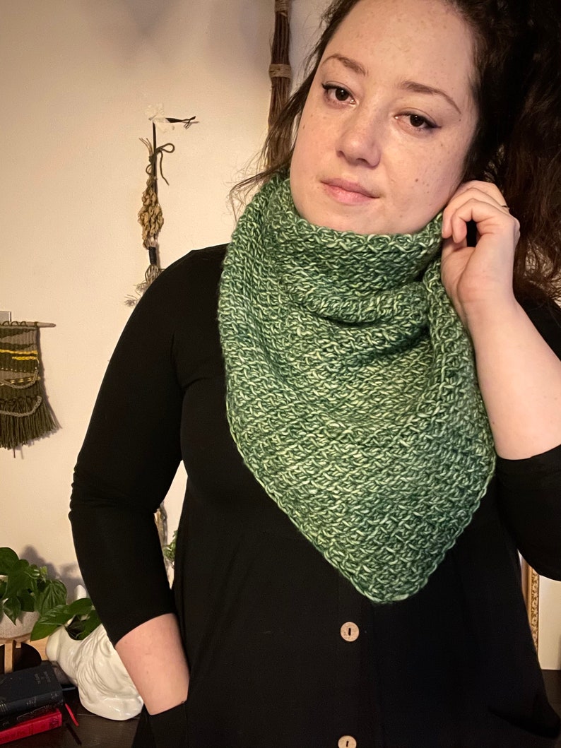 Brown haired woman in front of dried herbs with a sample of the Gabriel Tunisian crochet shawl pattern made using green ply yarn, display the unique texture of the beginner friendly easy crochet stitch project. Shown in kerchief size option reversed