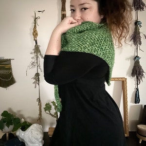 Brown haired woman turned to the side in front of dried herbs with a sample of the Gabriel Tunisian crochet shawl pattern made using green ply yarn, showing texture of the beginner friendly easy crochet stitch project. Shown in kerchief size option