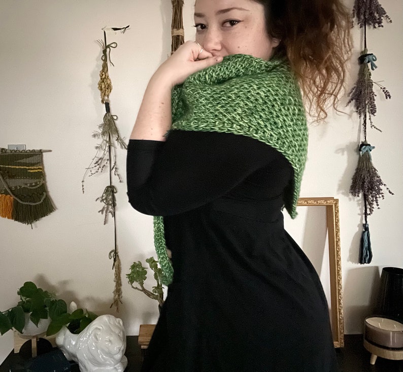 Brown haired woman turned to the side in front of dried herbs with a sample of the Gabriel Tunisian crochet shawl pattern made using green ply yarn, showing texture of the beginner friendly easy crochet stitch project. Shown in kerchief size option