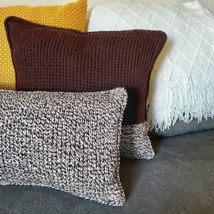 May include: Three knitted throw pillows in brown and white on a gray couch. The pillows are all different patterns and textures.