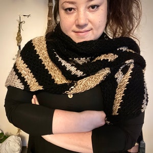 Brown haired woman with shawl tied over the shoulder that is a sample of the Gabriel Tunisian crochet shawl pattern made using black, silver and gold single ply yarn, display the unique texture of the beginner friendly easy crochet stitch project.