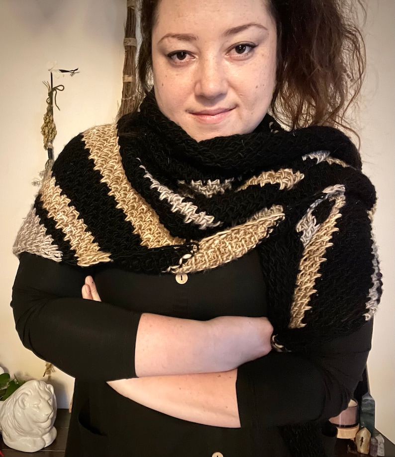 Brown haired woman with shawl tied over the shoulder that is a sample of the Gabriel Tunisian crochet shawl pattern made using black, silver and gold single ply yarn, display the unique texture of the beginner friendly easy crochet stitch project.