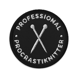 May include: A black circular patch with white embroidered text and crossed knitting needles. The text reads "PROFESSIONAL PROCRASKNITTER" around the edge. The knitting needles are white and detailed.
