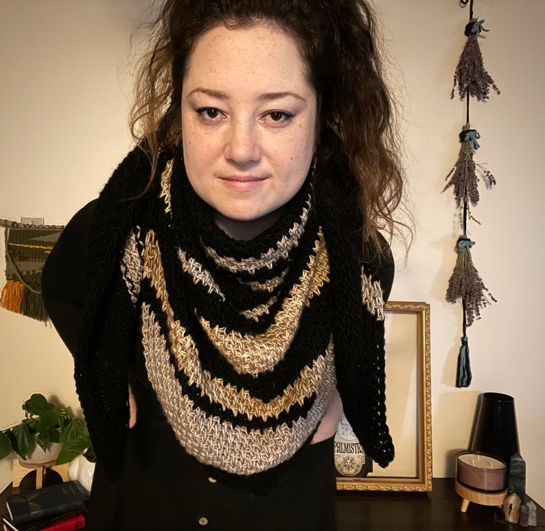 Brown haired woman in front of dried herbs with a sample of the Gabriel Tunisian crochet shawl pattern made using black, silver and gold single ply yarn, display the unique texture of the beginner friendly easy crochet stitch project.