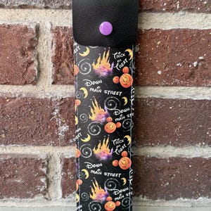 May include: A rectangular black sleeve with a flap secured by a purple snap button. The fabric features a repeating pattern of orange pumpkins, yellow crescent moons, and purple castle silhouettes with text reading Trick or Treat and Down Main Street.