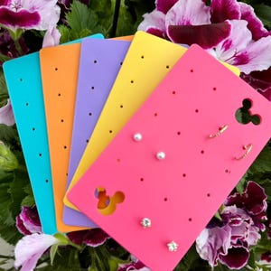 May include: A collection of colorful earring display cards in turquoise, orange, purple, yellow, and pink. The pink card features a Mickey Mouse cutout and holds several pairs of earrings, including studs and small hoops. The background is a floral arrangement.