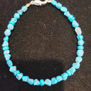 May include: A turquoise stone bracelet with a silver clasp. The bracelet is made of small, irregularly shaped, bright blue stones. The clasp is a simple, silver-colored lobster claw style.