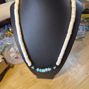 May include: A necklace with a strand of off-white, flat, circular beads. The necklace transitions to turquoise-colored, irregular-shaped beads, interspersed with small black beads. The necklace is displayed on a black mannequin.