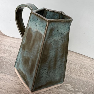 May include: A handmade ceramic jug with a distinctive angular form. The jug has a teal and brown glaze and a white base. The handle is curved and integrated into the design. A decorative and functional piece for serving drinks.
