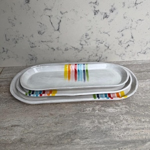 May include: A trio of oval ceramic serving platters. The white platters have a speckled texture and are decorated with vertical stripes in rainbow colours. The platters are stacked on top of each other.