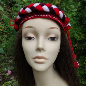 May include: A red, white, and burgundy braided headband with tassels. The headband is made of yarn and has a woven design.