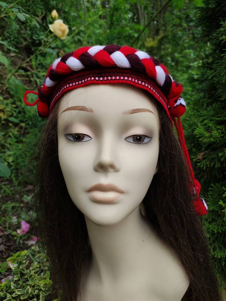 May include: A red, white, and burgundy braided headband with tassels. The headband is made of yarn and has a woven design.