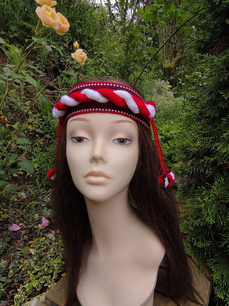 May include: A red and white braided headband with white pom-poms on the ends. The headband is worn on a mannequin with long brown hair.