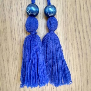 May include: Two blue tassels with blue and silver beads at the top. The tassels are hanging from a wooden surface.