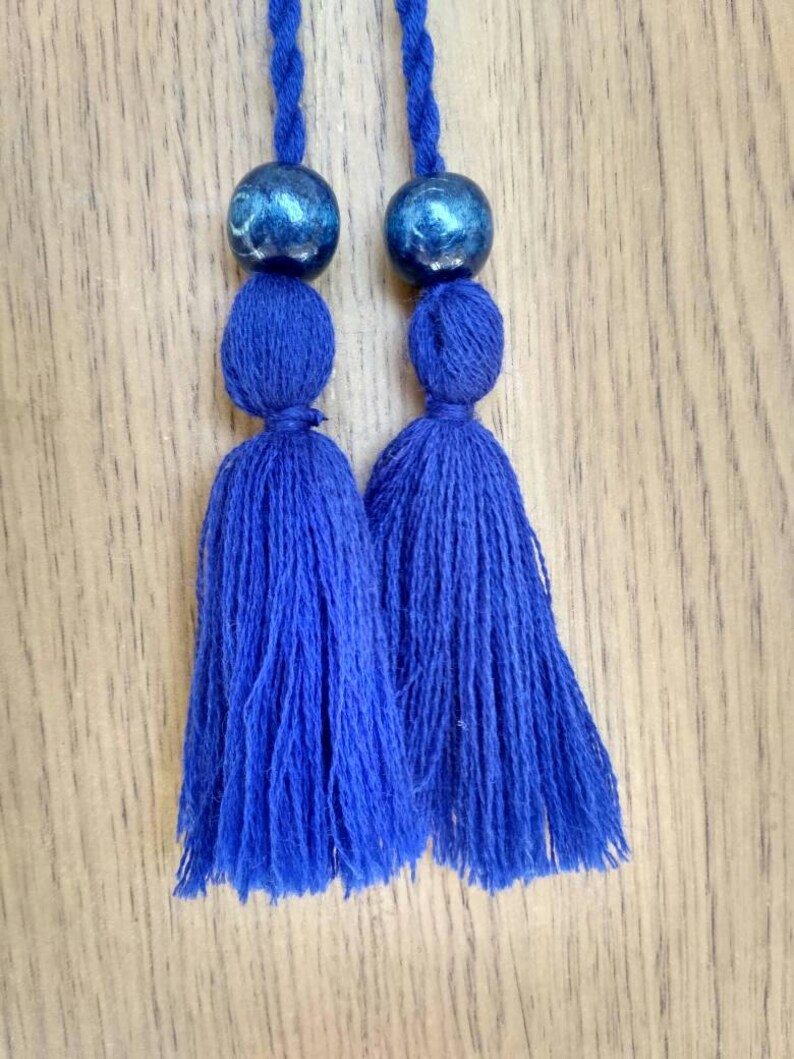 May include: Two blue tassels with blue and silver beads at the top. The tassels are hanging from a wooden surface.
